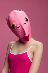 A woman wearing a pink fish head mask on Halloween stands and looks at the camera against a pink background. Conceptual art photo in a crazy way