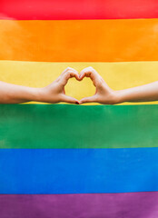 Close up of two woman's hands forming a heart with the LGBTQ rainbow flag in the background. Symbol for the homosexual community for pride month and the celebration day of sexual and love diversity