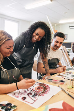 Smiling instructor assisting young woman in painting at desk in art class