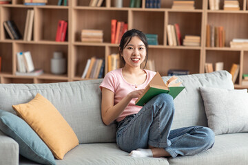 Domestic life concept. Positive asian lady holding paper book, sitting on comfy couch at home alone, free space