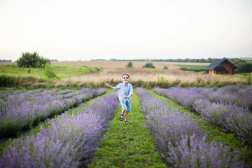 Smiling little boy running on the lavender field. Funny child in glasses dressed in a blue shirt and a straw hat running on the grass between lavender flowers. Horizontal photo