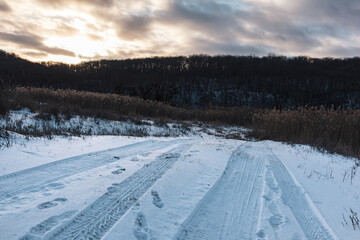 Fototapeta premium Snowy white rural road with tires prints in countryside. Evening winter landscape with sun in epic heavy clouds sky, reeds and dark trees forest hill