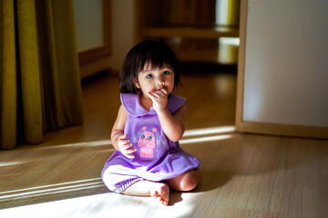 Portrait of Baby Girl wear Purple Dress sitting on the Floor and put her hand on her mouth in Empty room with reflection of light and shadow. Light and Shadow Concept. Looking at Camera.