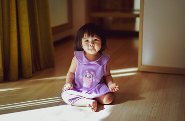 Portrait of Baby Girl wear Purple Dress sitting on the Floor and smiling to the camera in Empty room with reflection of light and shadow. Light and Shadow Concept. Looking at Camera.