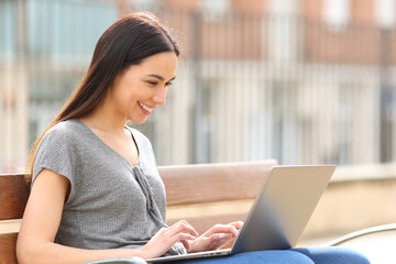 Happy woman using laptop sitting on bench in the street