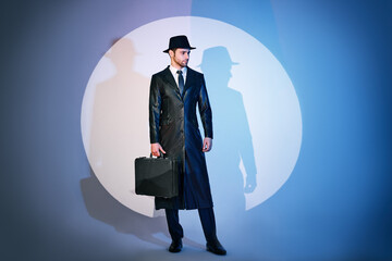 Full length portrait of handsome man in black coat and hat holding briefcase posing in the spotlight on studio background - obrazy, fototapety, plakaty
