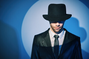 Portrait of handsome man in black coat hiding face behind his hat in the spotlight on studio background