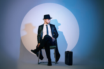 Handsome private detective sitting on chair in the spotlight on studio background - obrazy, fototapety, plakaty