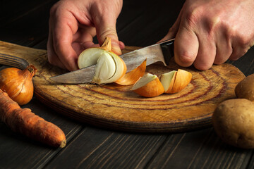 The chef cuts the onion with a knife on a wooden chopping board. Cooking vegetable dinner in the kitchen