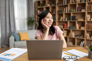 Portrait of happy dreamy asian lady sitting at desk with laptop at home, looking away, thinking and smiling, free space