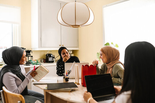 Cheerful young women wearing hijabs sitting at table in kitchen