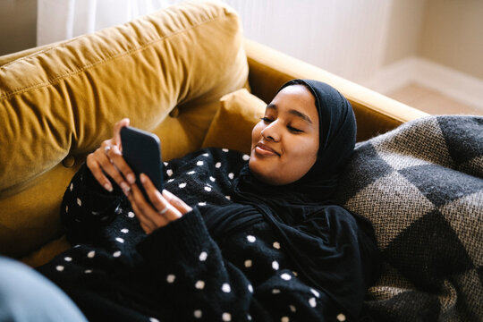 Smiling woman wearing hijab using mobile phone lying on sofa at home
