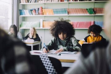 Boy with Afro hairstyle writing in book while sitting at desk in classroom