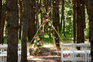 Fototapeta na wymiar Beautiful bohemian tipi arch decoration on outdoor wedding ceremony venue in pine forest with cones. Chairs, floristic flower compositions of roses, carpet, string fairy lights. Summer rural wedding.