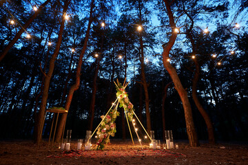 Festive string lights illumination on boho tipi arch decor on outdoor wedding ceremony venue in pine forest at night. Vintage string lights bulb garlands shining above chairs at summer rural wedding. - obrazy, fototapety, plakaty