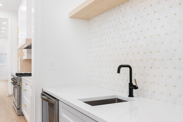 Sink detail shot with a black and gold faucet, white cabinet and marble countertop, and a white and gold mosaic tile looking out towards a modern kitchen. - obrazy, fototapety, plakaty