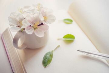 Bouquet of blooming apple flowers in a small mug vase on a creamy blank book with a pen and green leaves