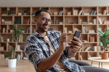 Happy arab young man sitting in chair with cellphone, chatting with friends, resting at home interior, free space