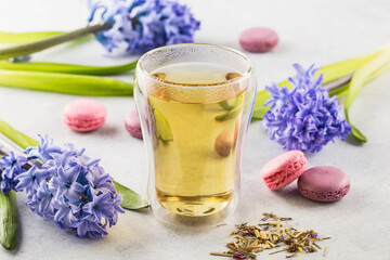 Hot lemongrass tea in a glass with double walls, dried lemongrass tea and macaroons on a light background decoraated with flowers .