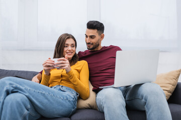 Cheerful woman holding cup of coffee near arabian boyfriend with laptop on couch.