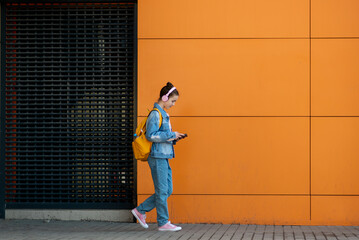 Cute teenage girl with a gadget and headphones. Against the background of an orange wall. The concept of modern youth. Kids with gadgets. Modern world. Lifestyle. Minimalism.