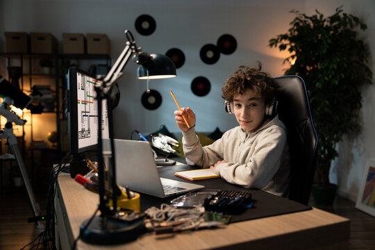 A young boy sits at his desk with wireless headphones in front of a computer taking notes with a pencil, drawing a design, passionate about architecture