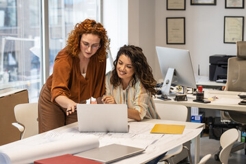 Two female coworkers are trying to solve a problem of a new project.They were standing by the desk and looking at the blueprint.Working day at the office.	
