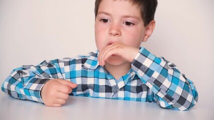 A brooding 4-year-old boy in a plaid blue shirt bites his nails while sitting at a table on a white background. Bad habits of children
