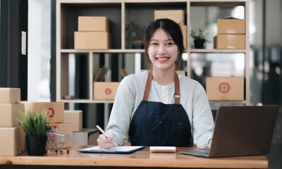 A portrait of a young Asian woman, e-commerce employee sitting in the office full of packages in the background write note of orders and a calculator, for SME business ecommerce and delivery business.