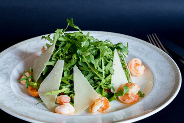 Arugula with shrimps and parmesan on a white plate on dark table