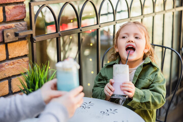child with open mouth holding glass of milk dessert near blurred mom in street cafe.