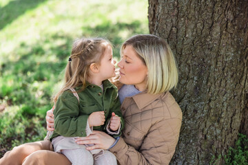 woman and girl in stylish clothes sitting under tree in park and looking at each other.