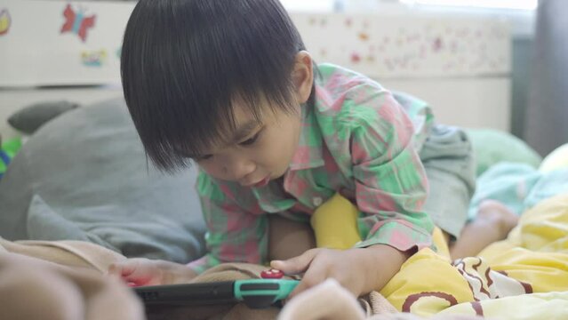 Little boy in a shirt wear lying in bed and playing a video game on portable mobile game or mobile phone while spending free time at home