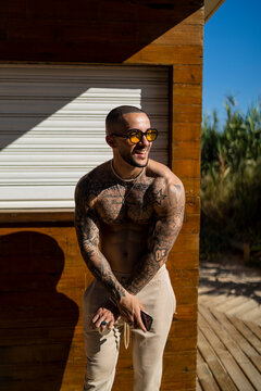 Chico joven rapado con tatuajes y musculado posando con ropa actual en la calle con mucho sol