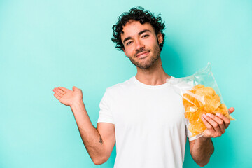 Young caucasian man holding a bag of chips isolated on blue background showing a copy space on a palm and holding another hand on waist.