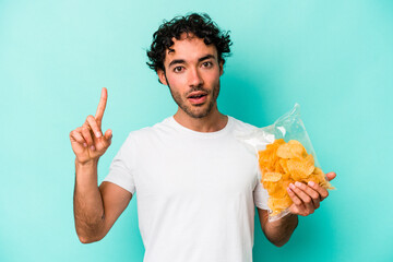 Young caucasian man holding a bag of chips isolated on blue background having an idea, inspiration concept.