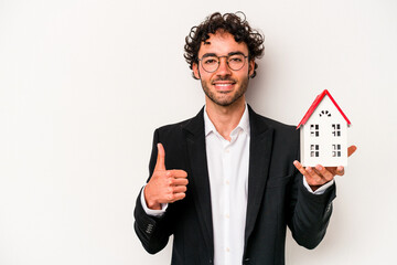 Young caucasian business man holding a toy house isolated on white background smiling and raising...