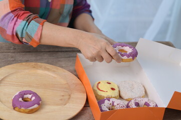 The woman's right hand was taking out a 1 purple glazed donut from an orange donut box and placing...