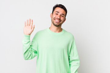young handsome hicpanic man smiling happily and cheerfully, waving hand, welcoming and greeting...
