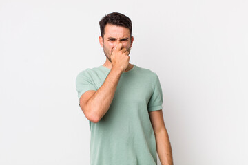 young handsome hicpanic man feeling disgusted, holding nose to avoid smelling a foul and unpleasant...