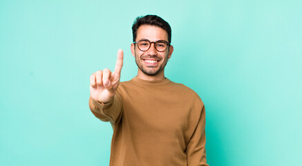 young handsome hicpanic man smiling and looking friendly, showing number one or first with hand...