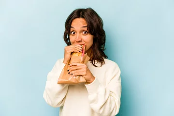 Poster Young hispanic woman eating a sandwich isolates on blue background © Asier