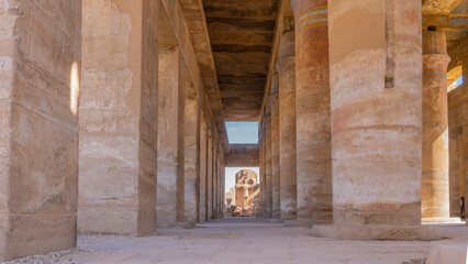 Smooth rows of columns in the Karnak temple of Luxor. Ahead, in the gap between the columns, a dilapidated statue is visible against the blue sky. Egypt