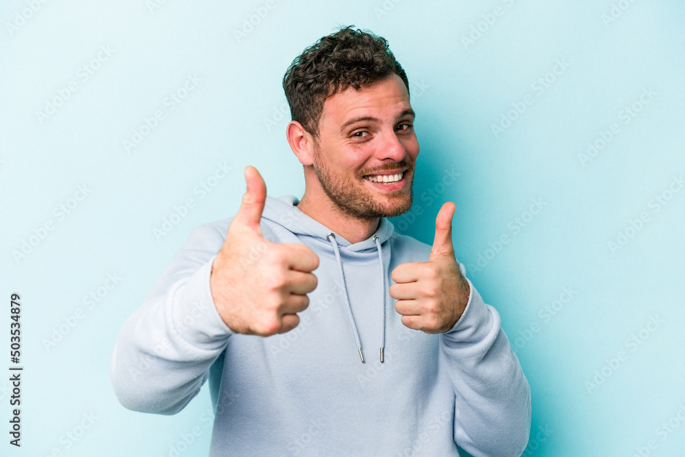 Wall mural young caucasian man isolated on blue background raising both thumbs up, smiling and confident.