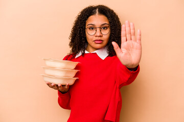 Young African American woman holding tupperware isolated on beige background standing with...