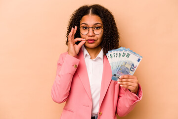 Young African American business woman holding banknotes isolated on beige background with fingers on lips keeping a secret.