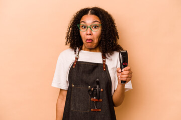 Young African American hairdresser woman isolated on beige background shrugs shoulders and open eyes confused.