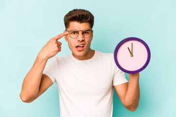 Young caucasian man holding a clock isolated on blue background showing a disappointment gesture with forefinger.