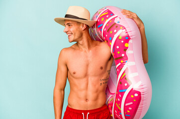 Young caucasian man holding an inflatable donut isolated on blue background looks aside smiling, cheerful and pleasant.