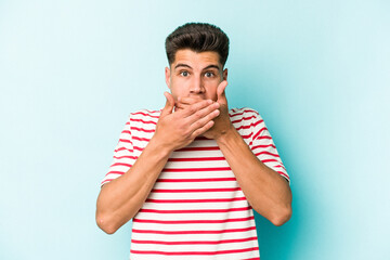 Young caucasian man isolated on blue background covering mouth with hands looking worried.